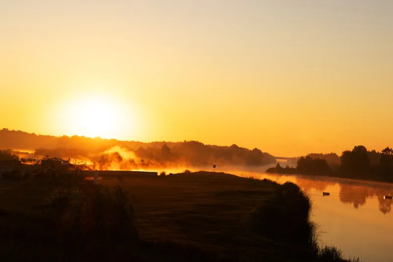 A golden sunrise over a wide, still river with mist rising off the water and dark trees along the far bank