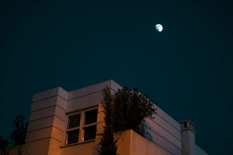 A house at night beneath a deep blue sky with the moon overhead, one upstairs window glowing warm from a lamp still on inside
